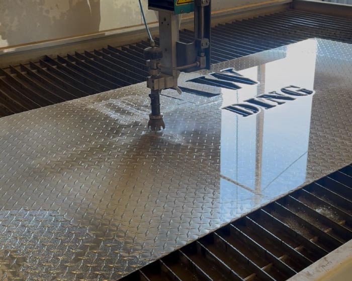 High-precision water jet machine cutting the RAW Welding logo into a polished aluminum diamond plate sheet, illustrating the capability to cut intricate branding and designs without heat distortion.