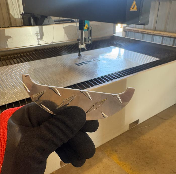 Close-up of a gloved hand holding a precision-cut aluminum diamond plate piece, showcasing the clean edges and intricate detail achieved by RAW Welding's state-of-the-art water jet cutting technology.