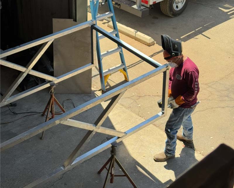 High-angle view of a welder in full PPE using a handheld grinder to finish the welds on a large, custom-fabricated aluminum frame structure in an outdoor work area