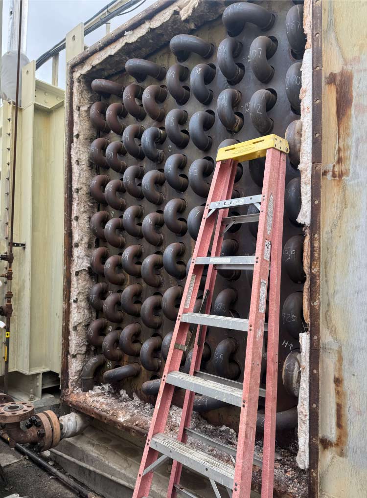 Large-scale industrial heat exchanger maintenance showing rows of carbon steel return bend tubing and a ladder positioned for inspection during a facility turnaround or outage