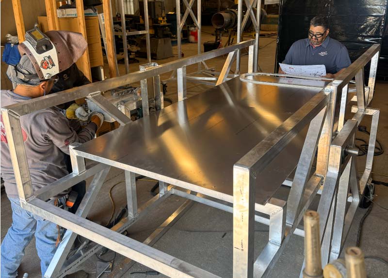 What Are Durable Metals? Two technicians in a metal fabrication shop working on a large custom aluminum structure; one wearing a welding helmet and welding a joint, while the other reviews detailed engineering blueprints