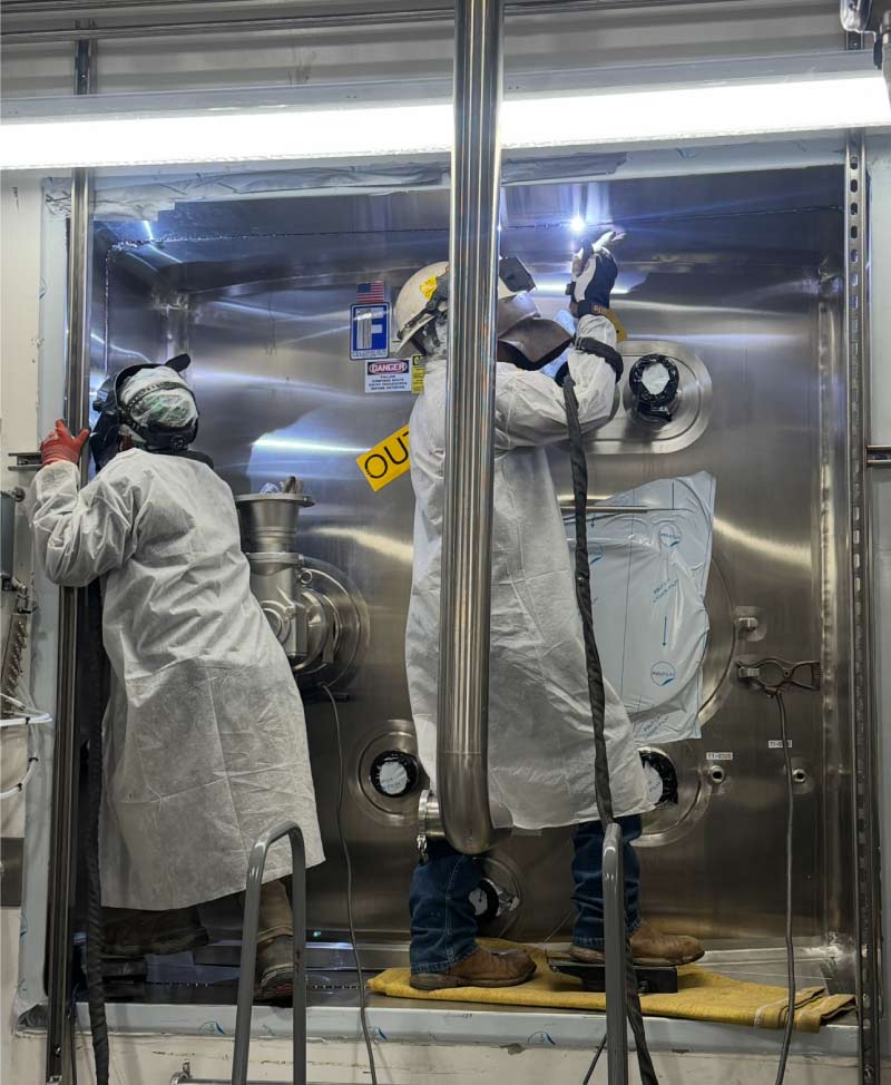 Heavy industrial pipe fitting with insulation and critical valve connection leading into a metal-sided refinery or processing plant wall.