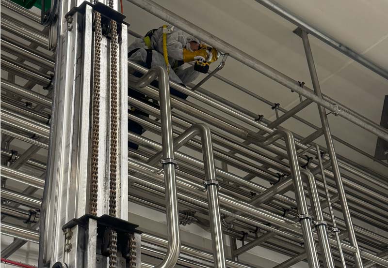 A technician wearing a white protective suit and safety harness working high up on a scissor lift to install or repair stainless steel industrial piping systems near the ceiling of a facility
