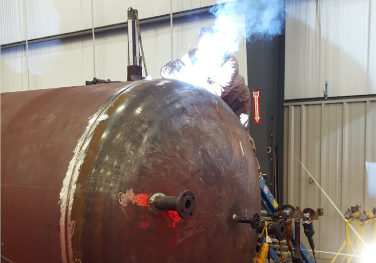 Skilled welder sitting atop a large industrial pressure vessel performing a structural repair weld with visible bright arc sparks and smoke fumes following Welding Safety Standards & Compliance 
