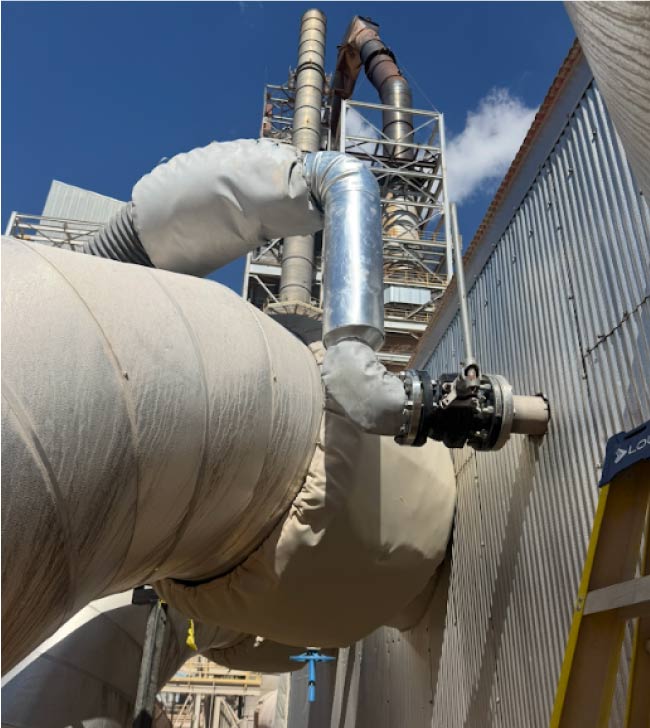 metal-experts Heavy industrial pipe fitting with insulation and critical valve connection leading into a metal-sided refinery or processing plant wall.