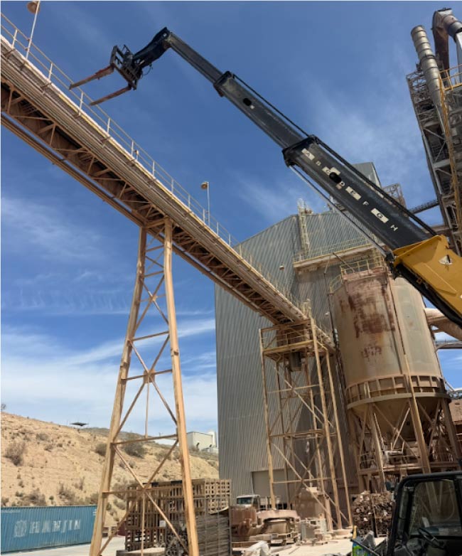 welding Telescopic handler lifting pipe onto a large industrial cement plant facility conveyor structure in the desert.