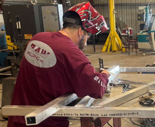 A RAW Welding Inc. specialist in a branded maroon long-sleeve shirt and protective helmet measuring metal fabrication components.
