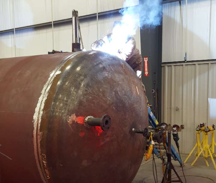 A skilled industrial welder performing precise arc welding on the end cap of a massive steel pressure vessel in a fabrication shop.