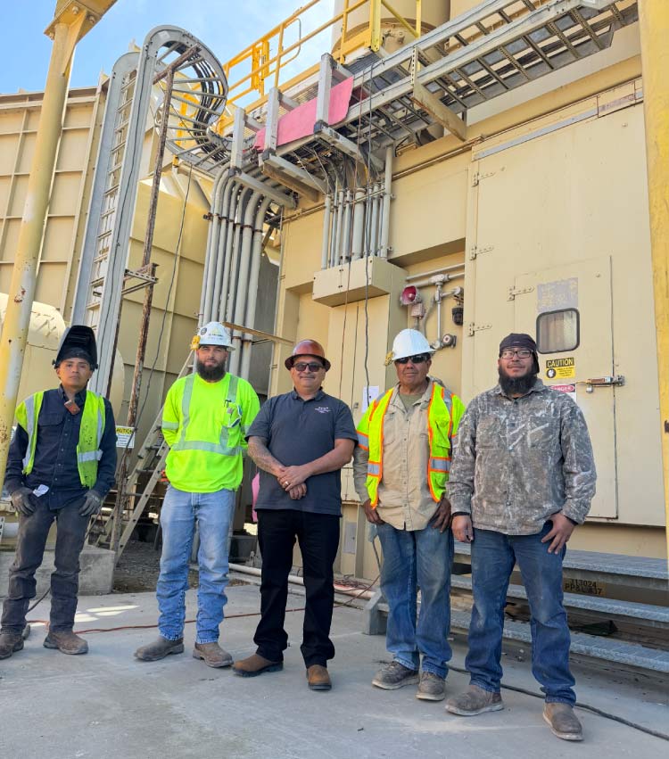A professional group photo of five RAW Welding Inc. team members standing in front of industrial machinery and complex piping. The team is wearing safety gear, including hard hats and high-visibility vests, representing their commitment to safety and expertise in industrial services.